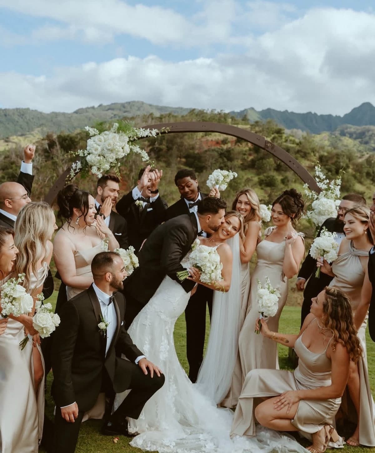 A wedding party laughing under a floral arch with mountains behind them